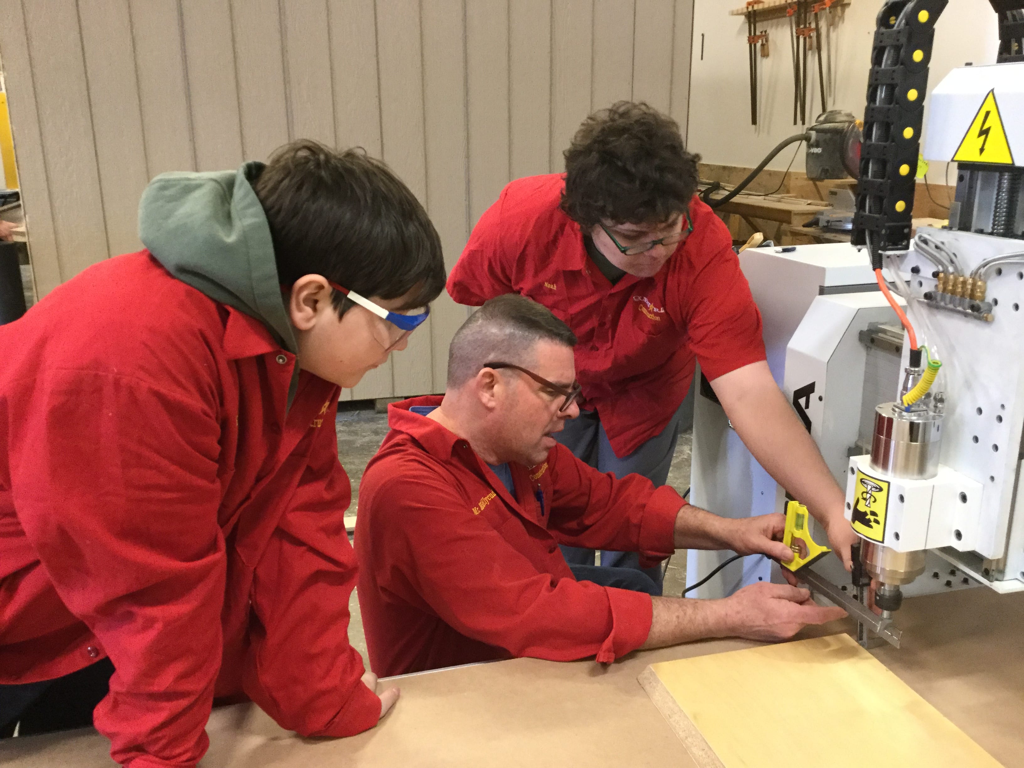 Noah and Colby learning CNC Image of three people in red workshirts working together on a CNC machine
