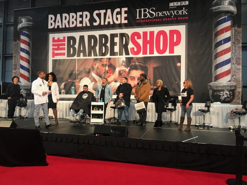 Image of men and women on stage with the words The Barber Shop and two barber poles.